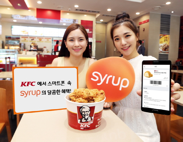 SK플래닛 시럽(syrup),전국 KFC 매장에서 O2O서비스 제공 홍보사진입니다. 왼쪽 모델은 KFC에서 스마트폰 속 시럽의 달콤한 혜택이라고 쓰여있는 배너를 들고 있고 오른쪽 모델은 시럽 앱 아이콘과 앱 구동화면을 들고 있습니다. 